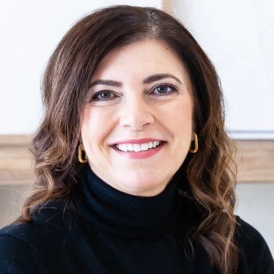 A woman with brown hair, wearing a black turtleneck and gold hoop earrings, smiling at the camera.