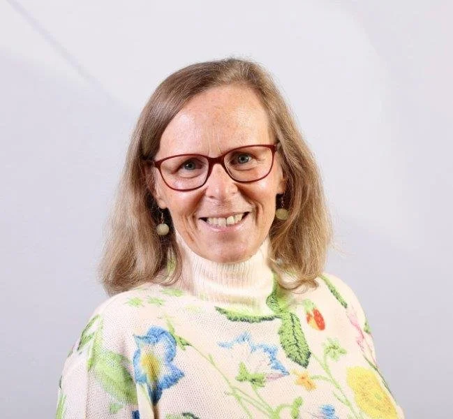 A woman with shoulder-length light brown hair, wearing glasses, earrings, a white turtleneck, and a cream cardigan with colorful floral embroidery, smiling at the camera.