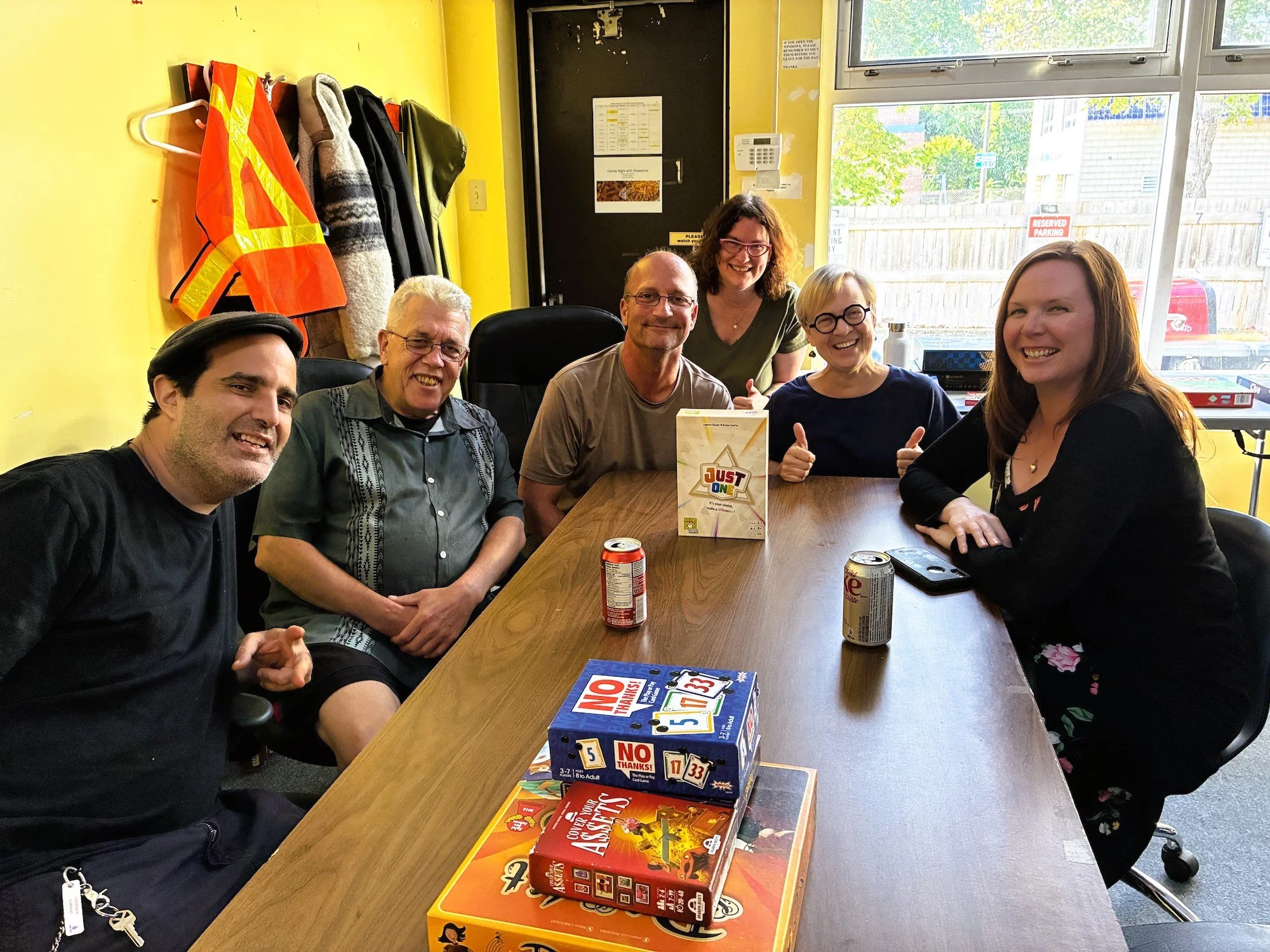 Group of six people sitting around a conference table, smiling at the camera, with board games and soda cans on the table inside a brightly lit room with windows and yellow walls.