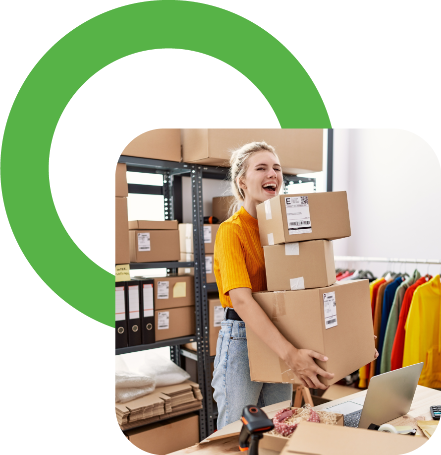 A young woman in a yellow shirt smiling and holding three cardboard shipping boxes in a storage or warehouse room with shelves of boxes and colorful clothing on racks.