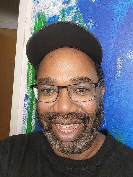 A man with glasses and a beard smiling, wearing a black cap and black shirt, standing in front of a colorful, abstract painted wall.