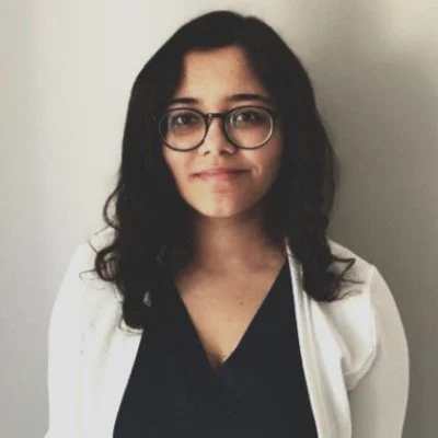 A woman with long dark hair, glasses, and a slight smile, wearing a black top and white jacket, standing against a plain wall.