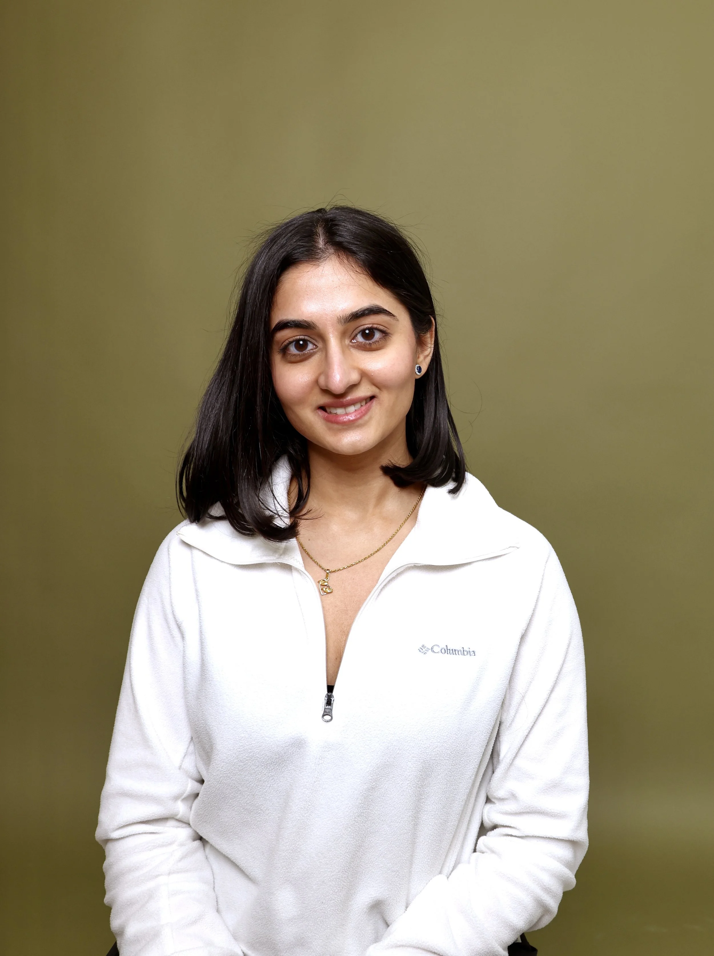 A young woman with dark hair, wearing a white Columbia zip-up jacket, smiling, standing against a plain olive green background.