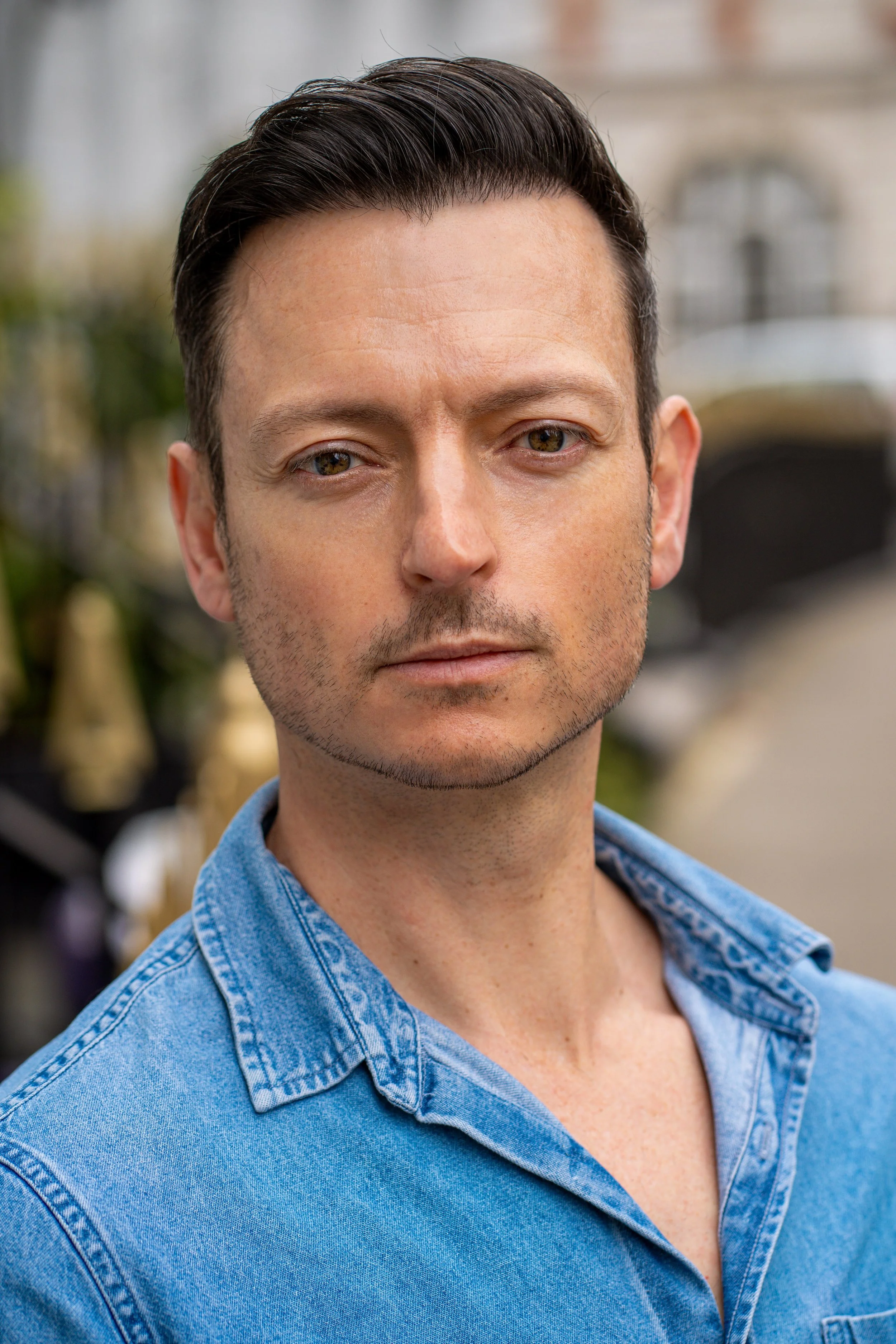 A man with dark, slicked-back hair and light facial hair, wearing a denim shirt, standing outdoors with blurred buildings and plants in the background.