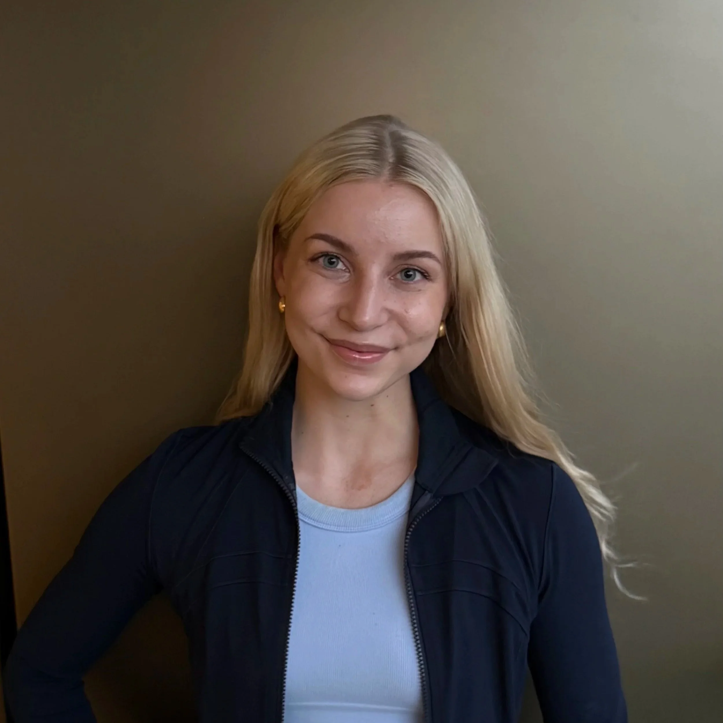 A young woman with long blonde hair, wearing a dark jacket over a light-colored shirt, standing against a neutral background, smiling with blue eyes and gold earrings.