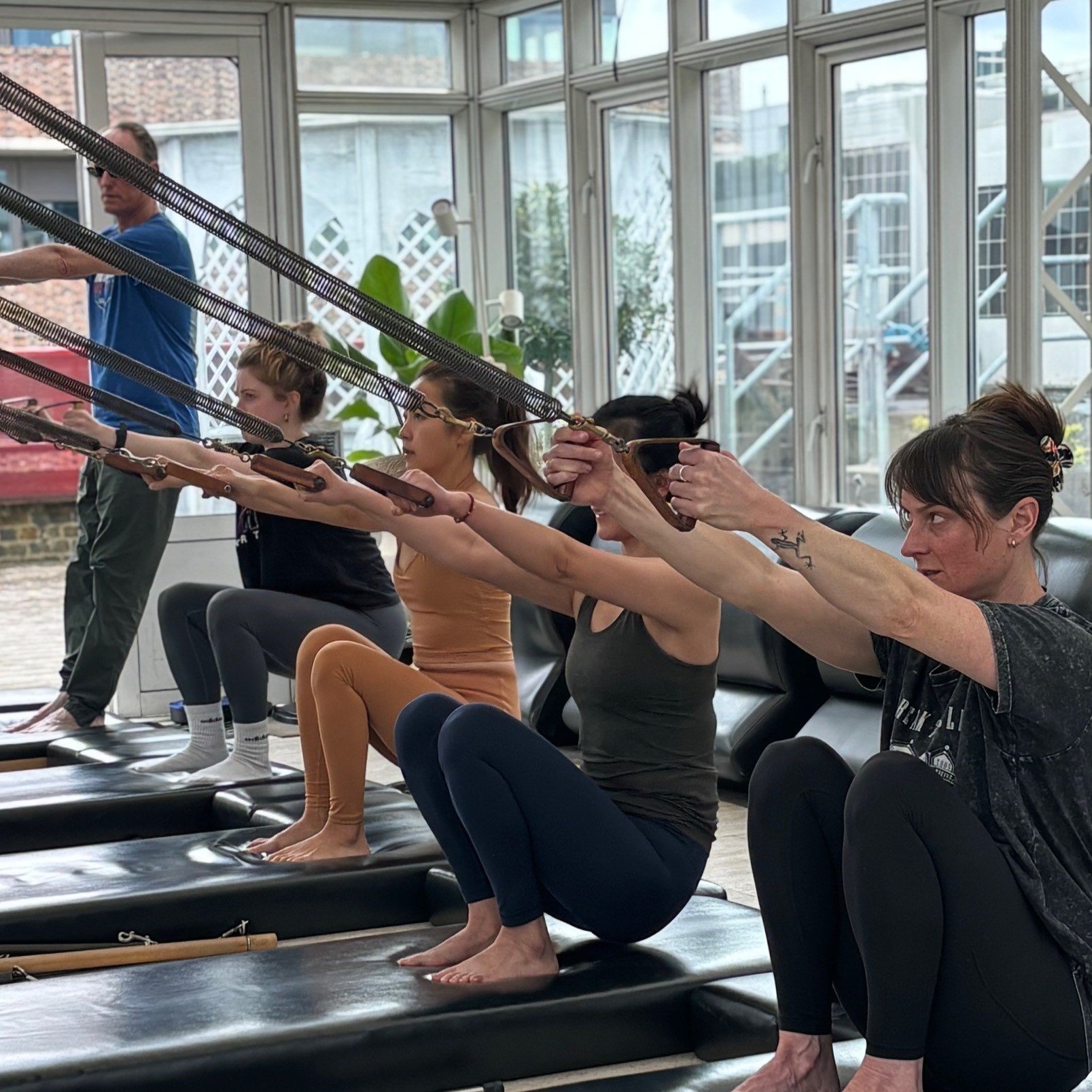 Women participating in a Pilates class, using resistance bands while sitting on black mats in a bright room with large windows.