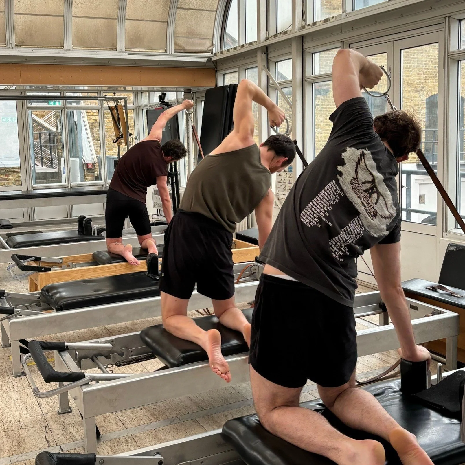 Three men performing Pilates exercises on reformer machines in a bright, urban Pilates studio with large windows and exposed brick walls.