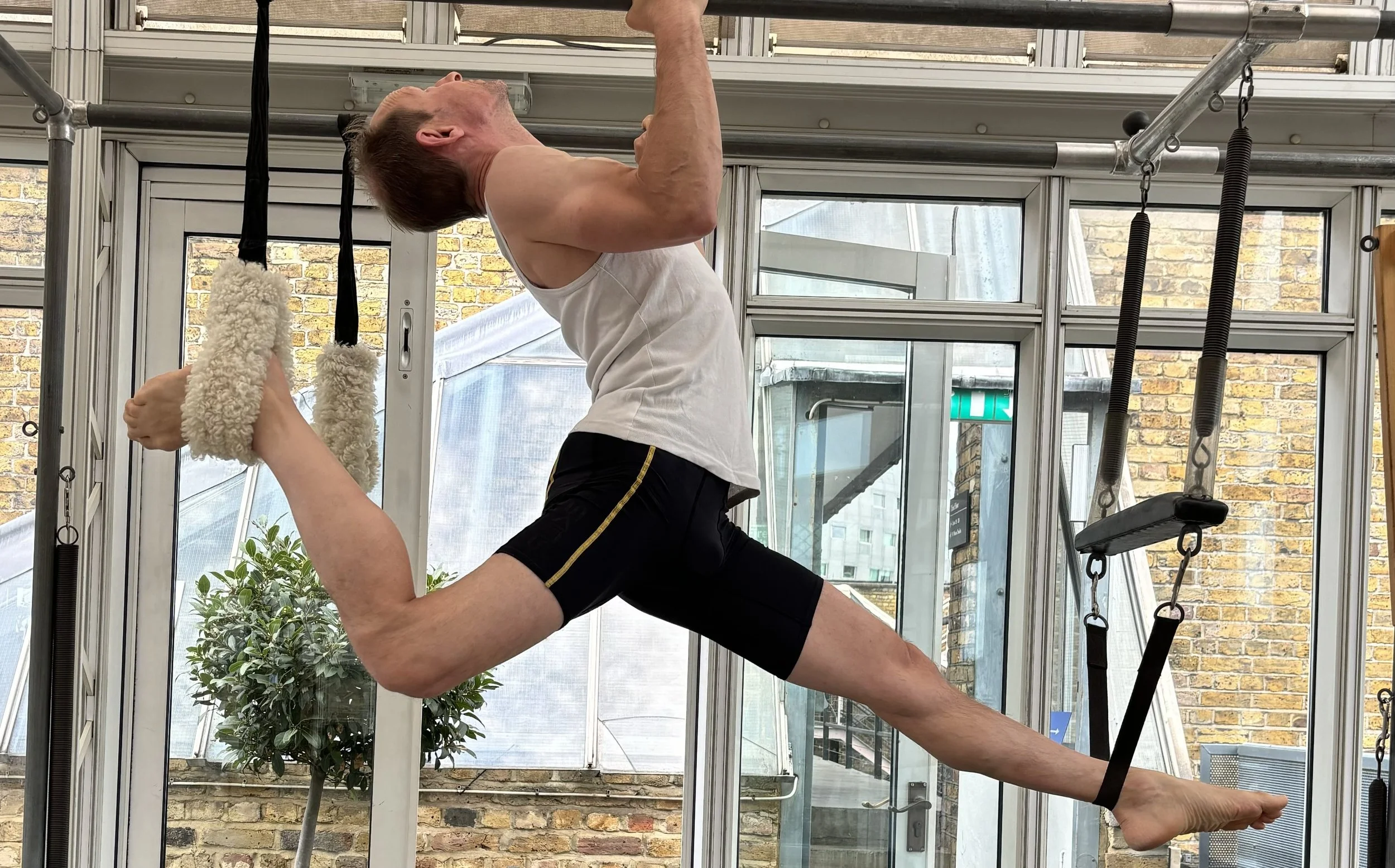 A man practicing aerial yoga or stretching in a glass-walled room, hanging from a suspension system with padded supports around his ankles, holding onto a bar with one arm, and extending his other arm. There are outdoor plants and brick buildings visible outside the windows.