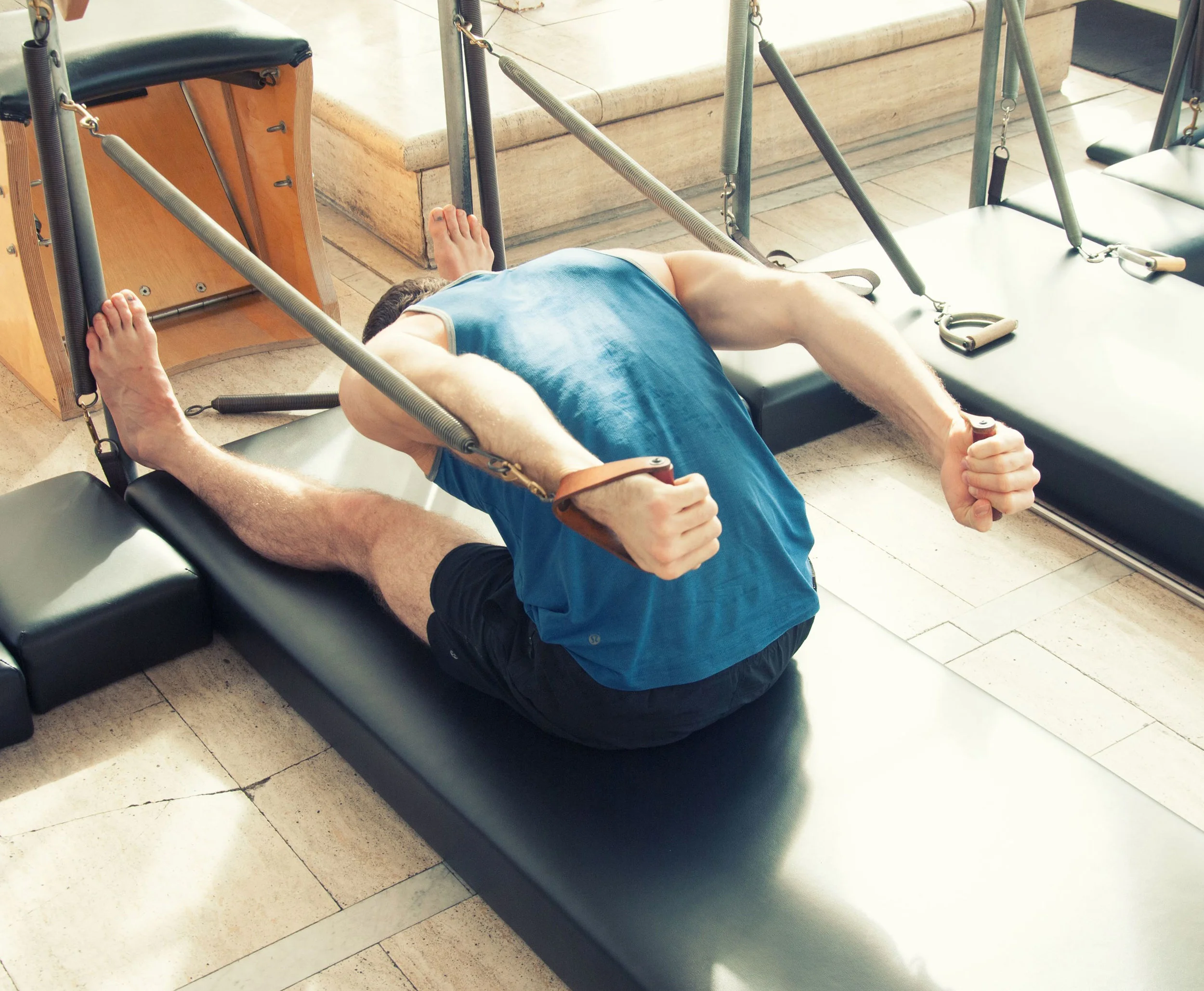 A man in a blue tank top doing a Pilates exercise on a reformer machine, with his upper body bent forward and holding handles, while his lower body rests on the bench.