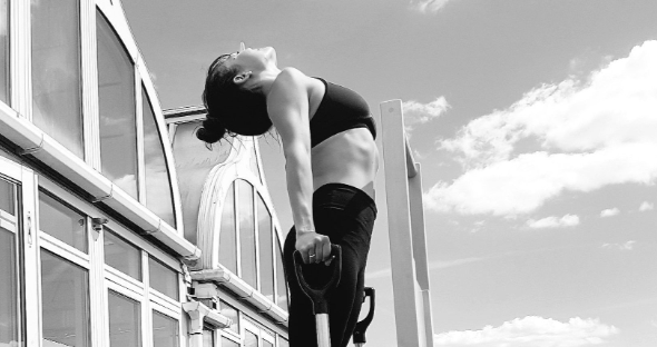 A woman in workout attire holding straps and leaning back outdoors near a glass building, with clouds and sky in the background.