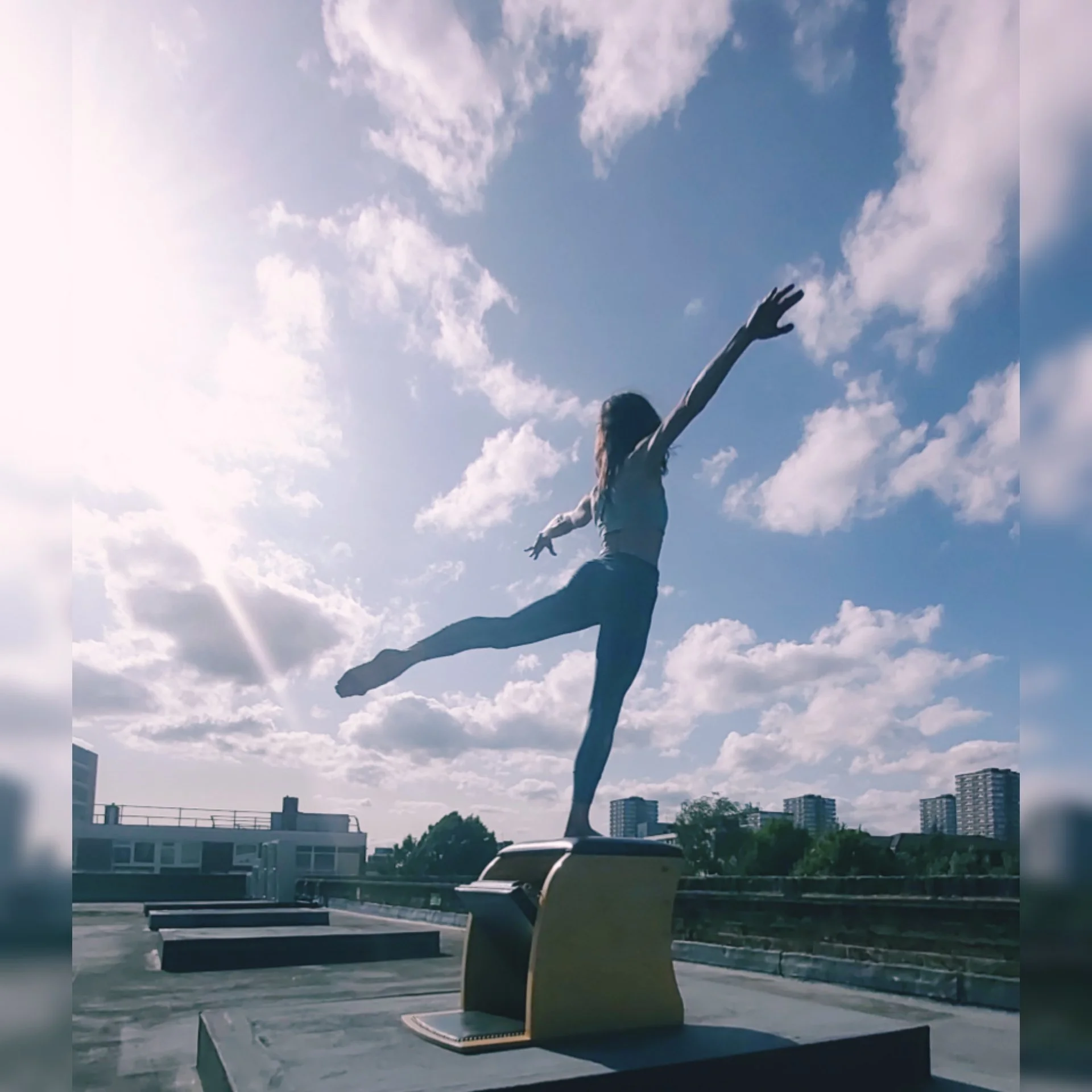 A woman standing on a rooftop air conditioning unit with her arms outstretched and one leg raised, mimicking a dance pose against a blue sky with scattered clouds.