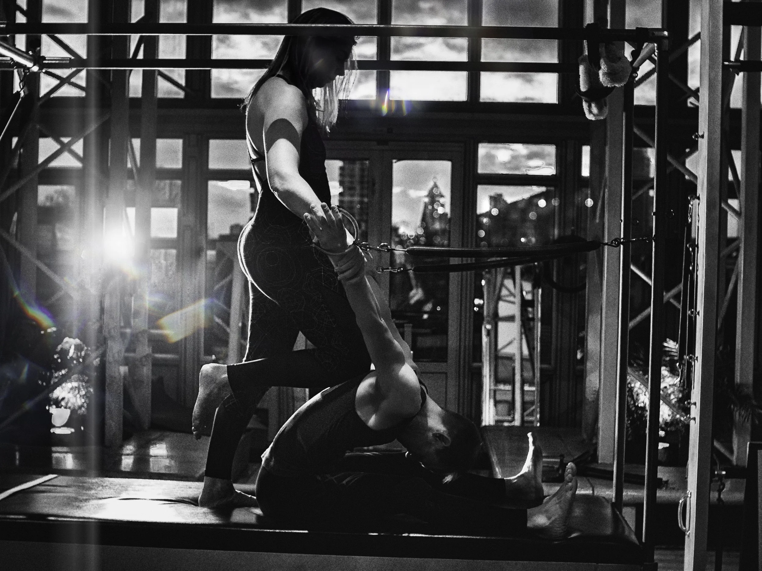 Two women practicing acro yoga inside a building with large windows and city skyline in the background; one is lying on the mat holding her leg up, while the other is supporting her positioned on top.