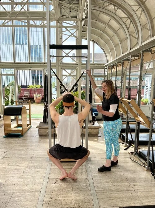 Person sitting on the floor in a fitness studio, wearing a white tank top and black shorts, facing away, with hands behind head, being instructed by a woman standing next to them. The woman has long hair, is wearing a black top and blue patterned pants. The studio has large windows and exercise equipment.