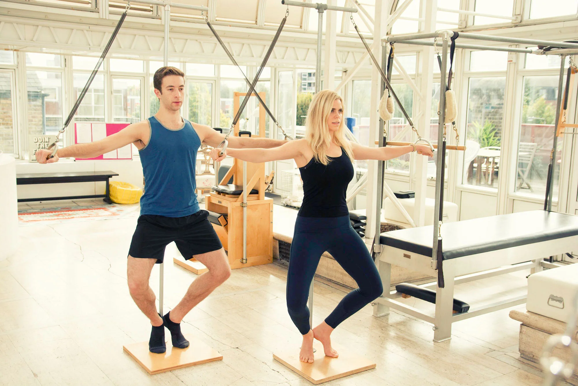 Two people exercising on Pilates reformers in a bright, sunlit room with large windows and various fitness equipment.
