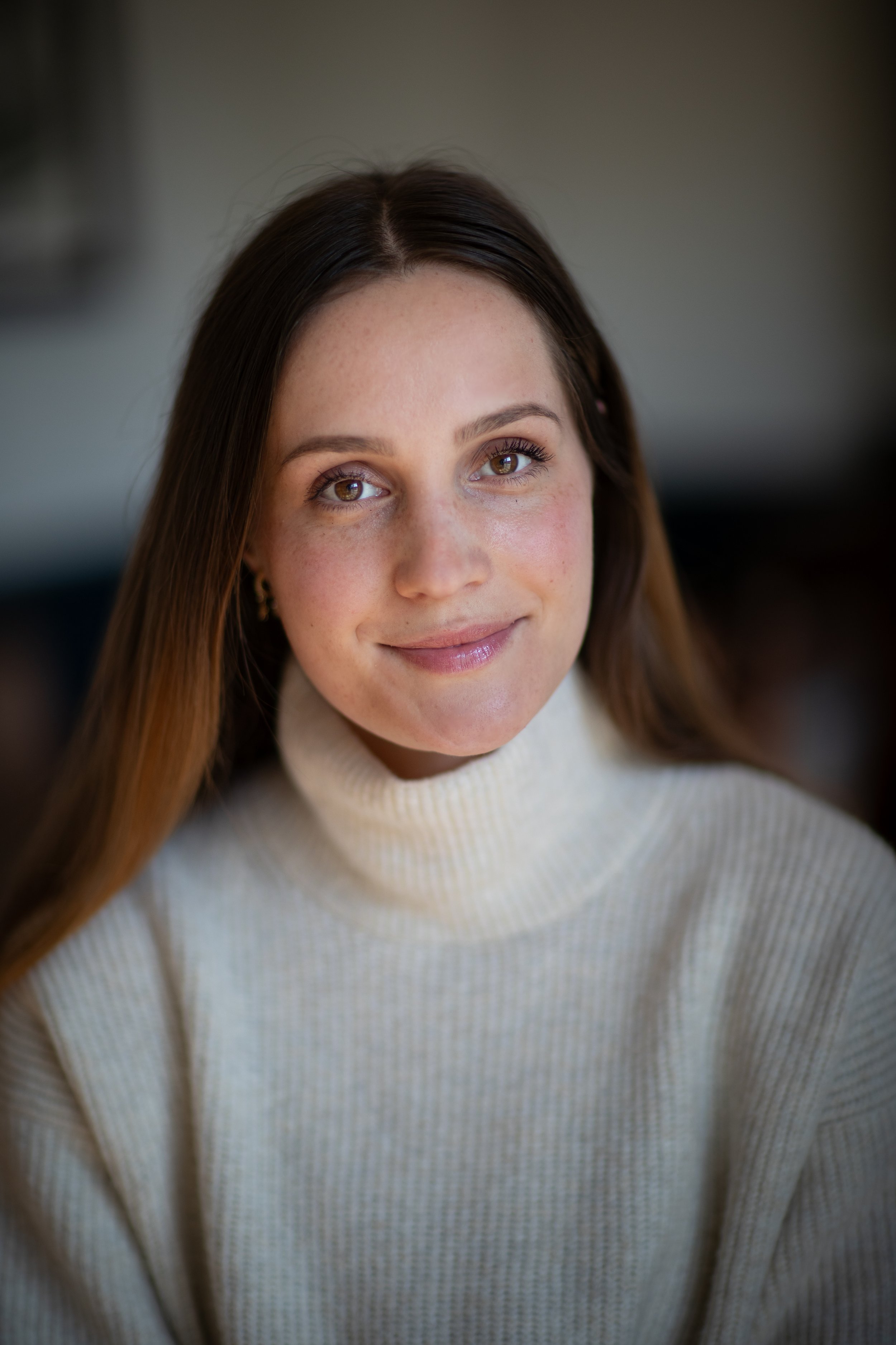 Close-up portrait of a young woman with long brown hair, hazel eyes, and a gentle smile, wearing a cream-colored turtleneck sweater.