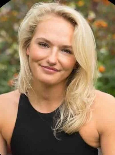 A blonde woman with wavy hair smiling at the camera, outdoors with blurred greenery and orange flowers in the background.
