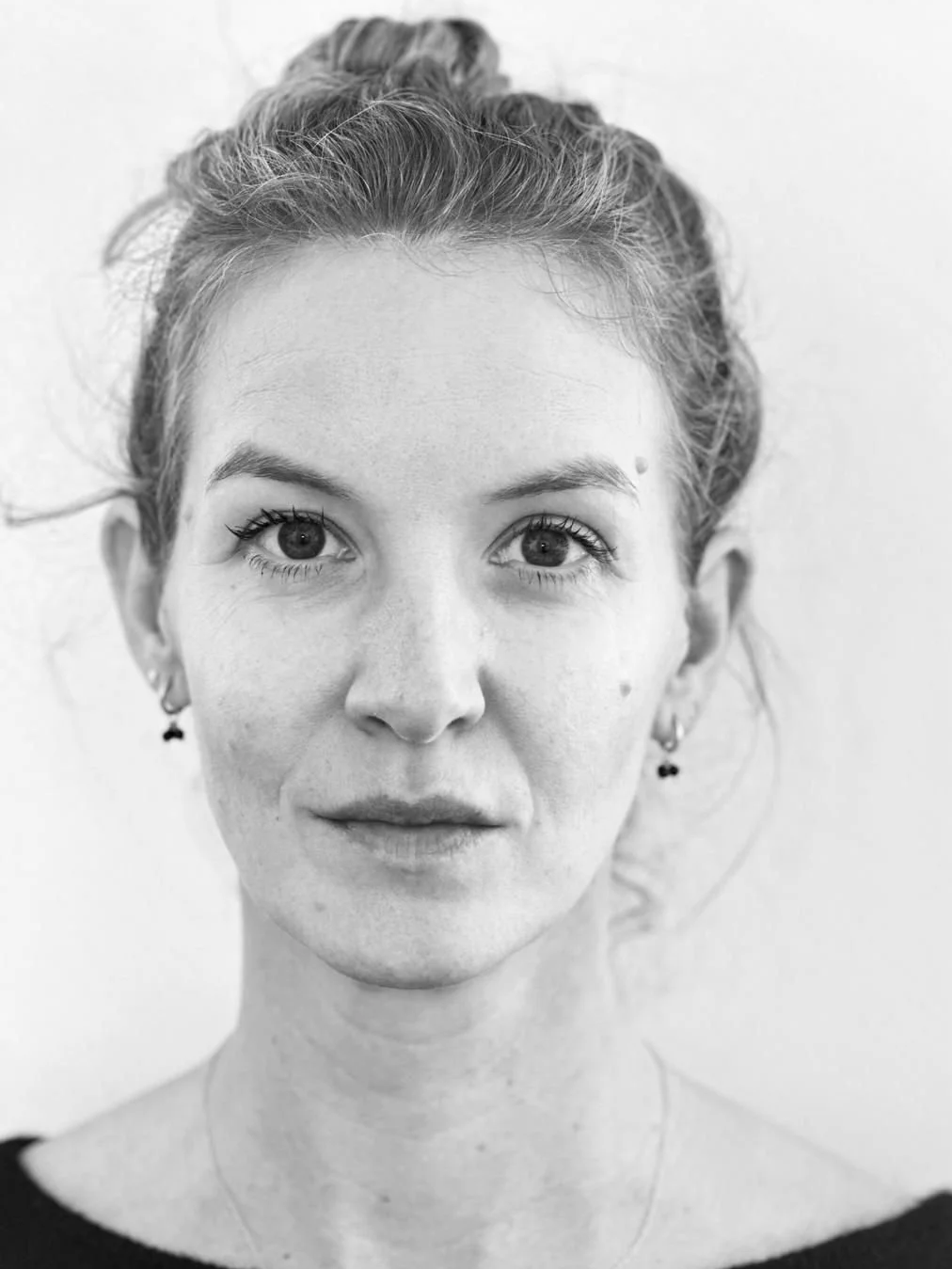Black and white portrait of a smiling woman with long hair, wearing earrings, outdoors near a body of water and a concrete wall.