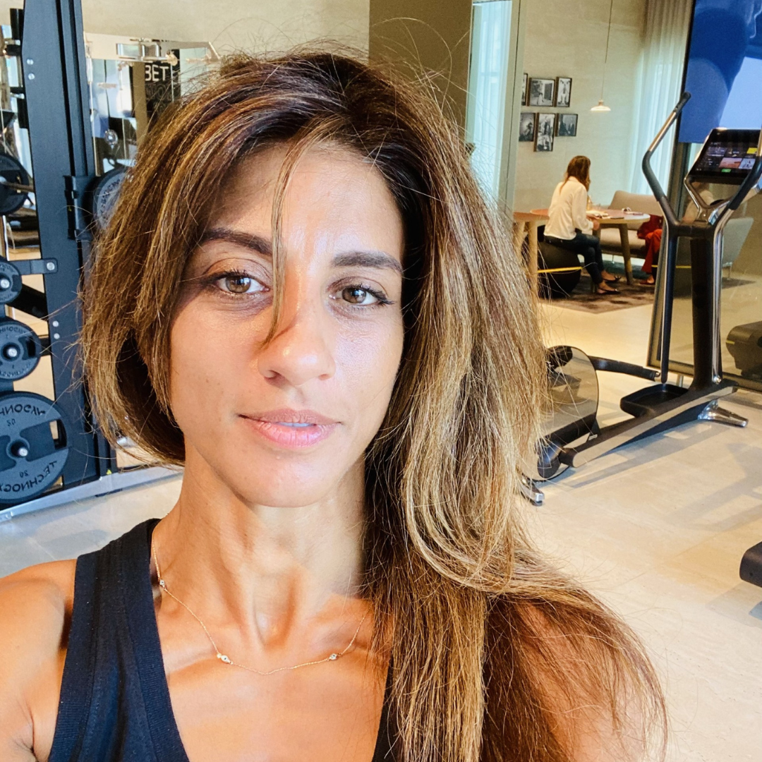 A woman with long brown hair smiling and sitting on a striped sofa in a room with white walls.