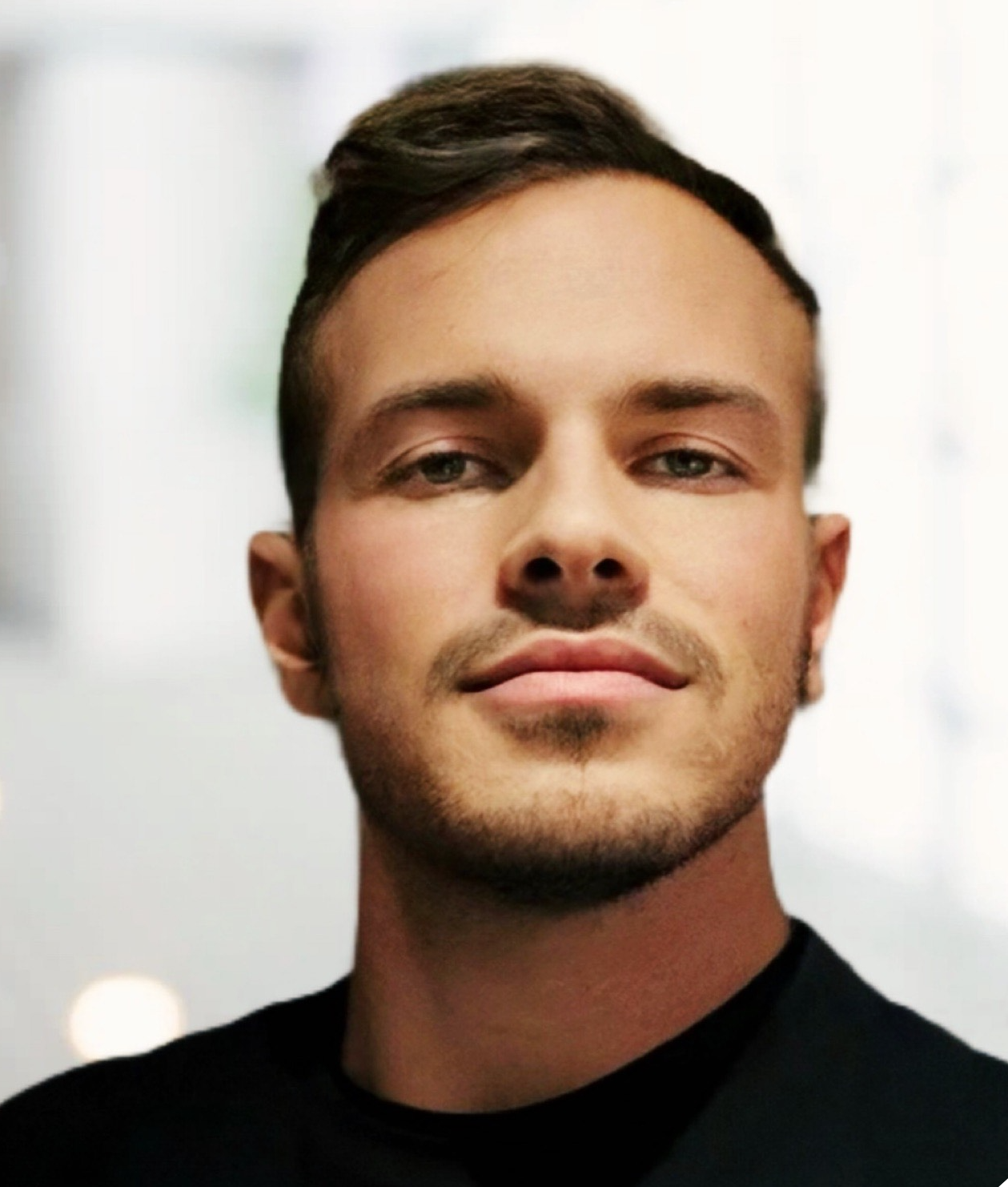 Portrait of a young man with short dark hair and slight smile, wearing a black top, in a bright, blurred indoor setting.