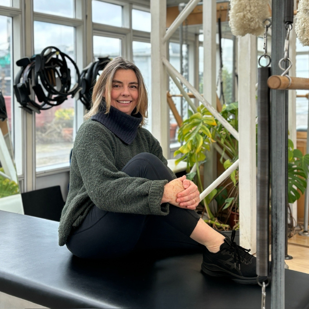 A woman in workout clothes doing a physical therapy exercise on a reformer machine, assisted by a woman with shoulder-length blonde hair wearing a green sweater, inside a fitness or physical therapy center.
