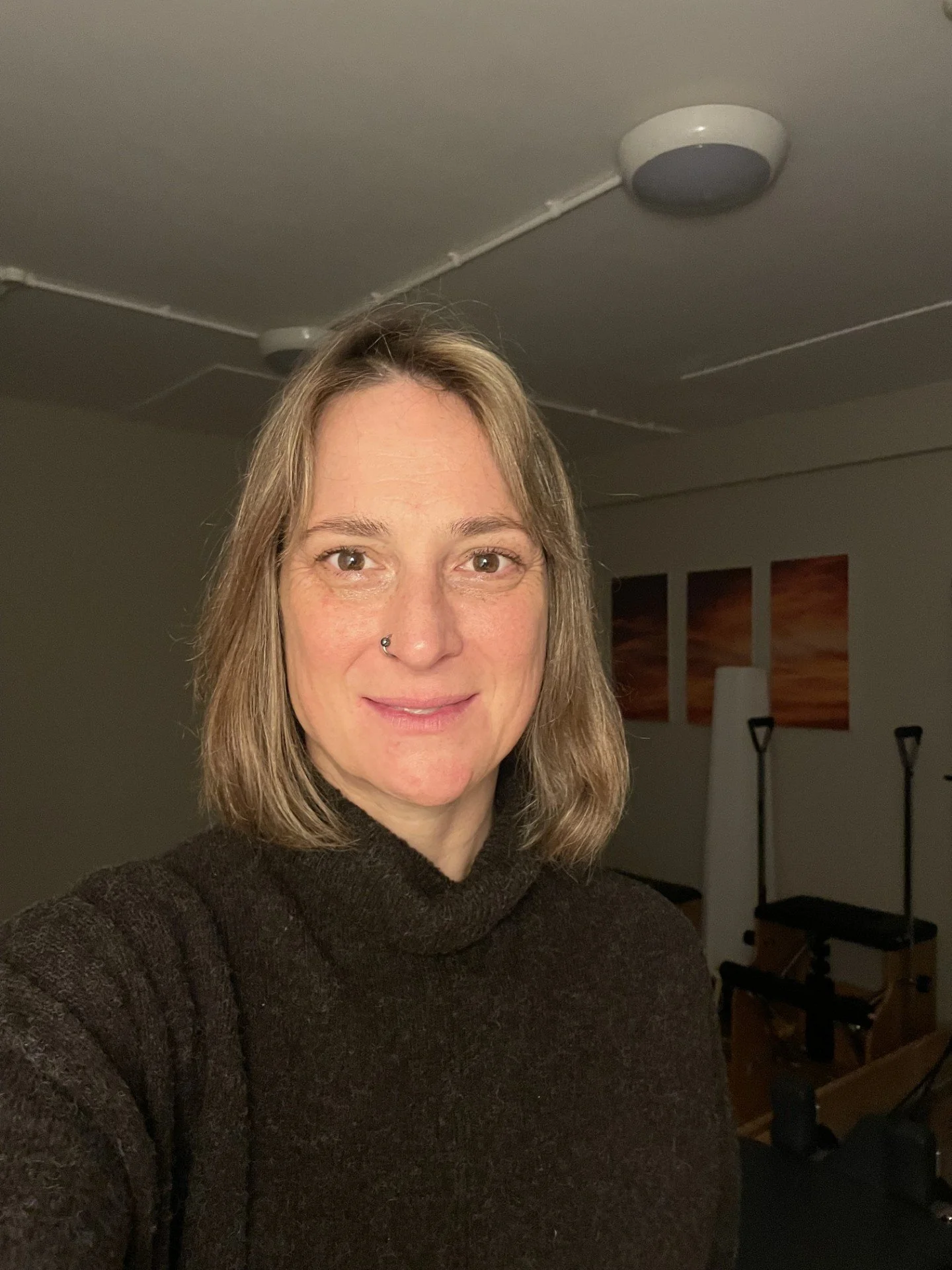 A woman with shoulder-length brown hair, smiling, wearing a black top with a strap visible on her shoulder, standing against a gray background.