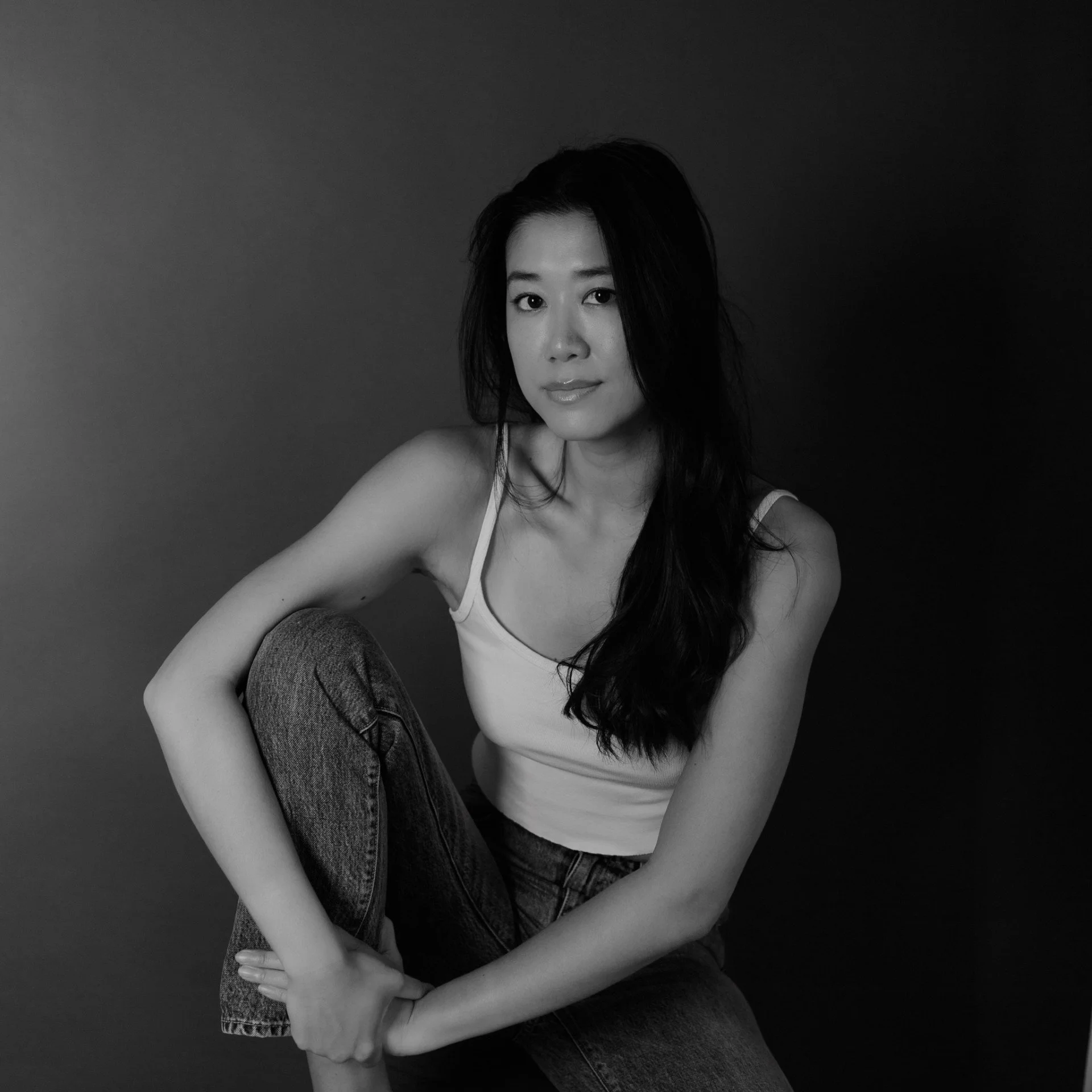 Black and white portrait of a young woman with long dark hair, sitting cross-legged, wearing a sleeveless top.