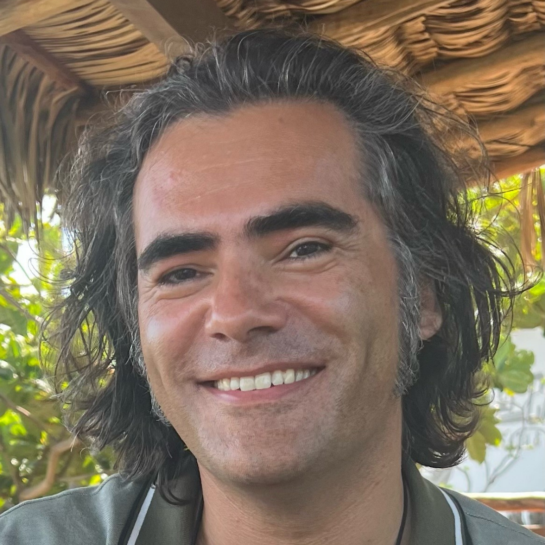 Smiling man with dark, curly, shoulder-length hair outdoors, with blurred green foliage in the background.