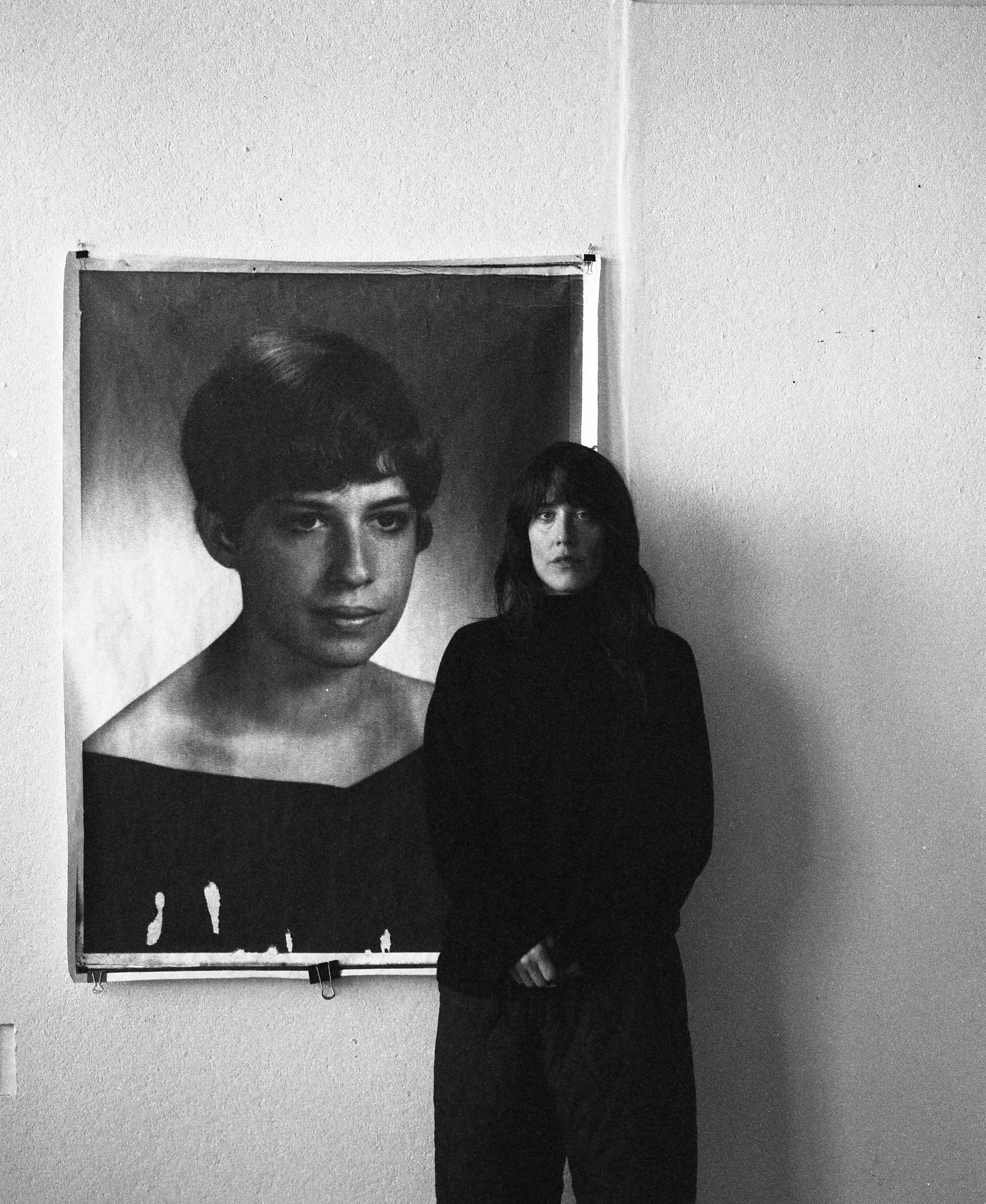 A woman with dark hair and a black outfit standing in front of a large black and white portrait of a woman with short hair, on a plain wall. Jae Matthews