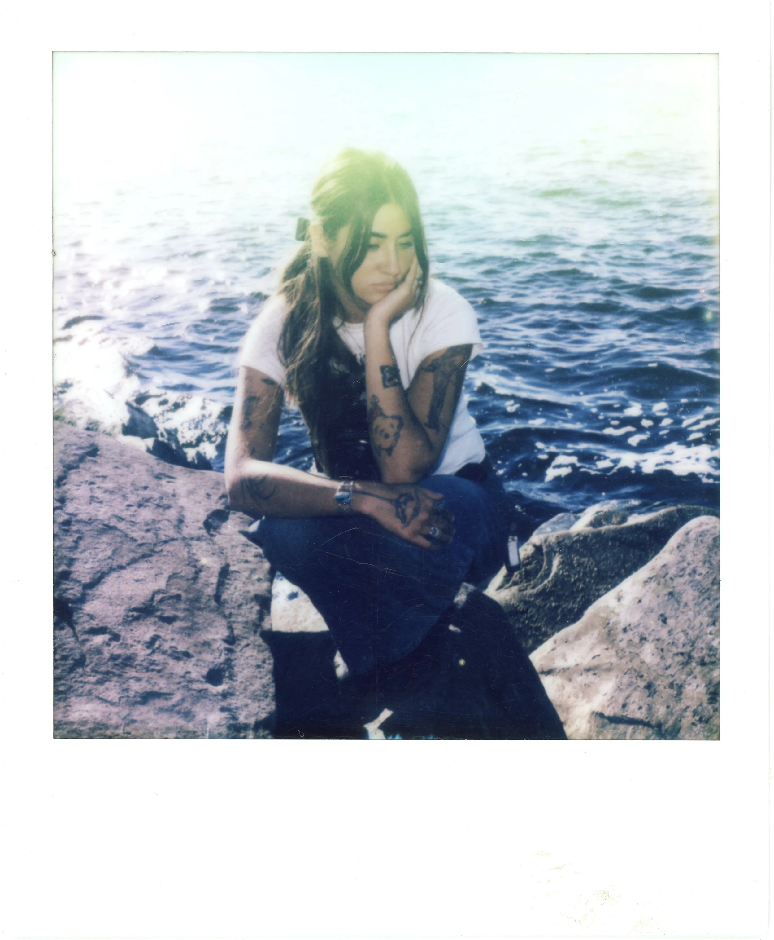 A young woman with tattoos sitting on rocks by the ocean, with waves visible in the background on a sunny day.