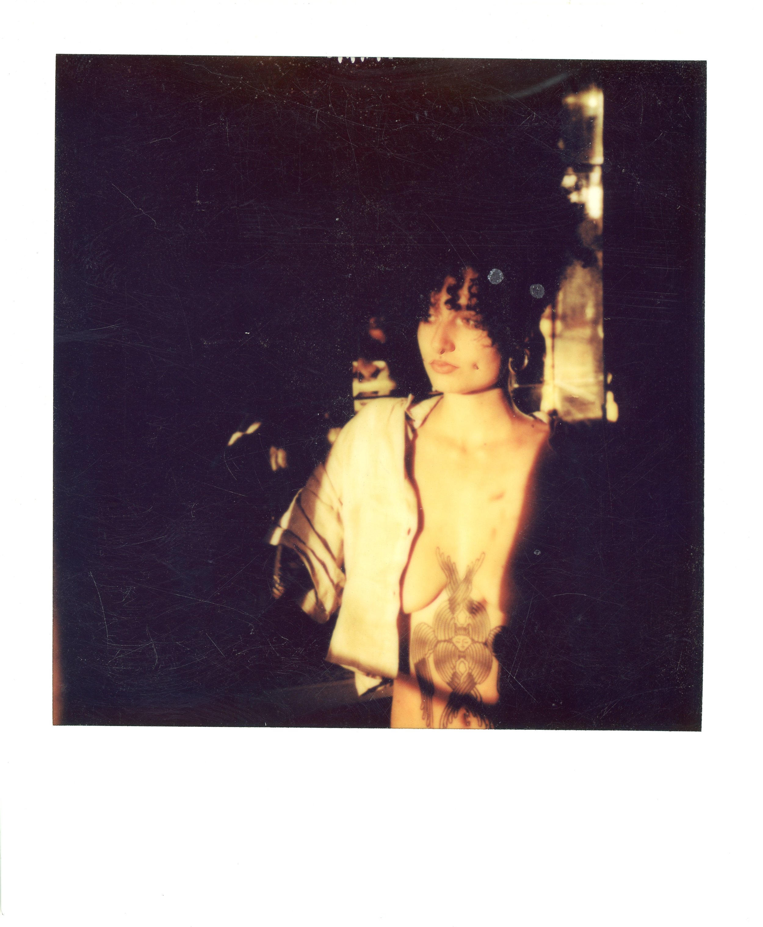 A woman with short dark hair and tattoos on her chest and arm, wearing a partially open shirt, stands in front of a window with sunlight casting shadows on her face and body.