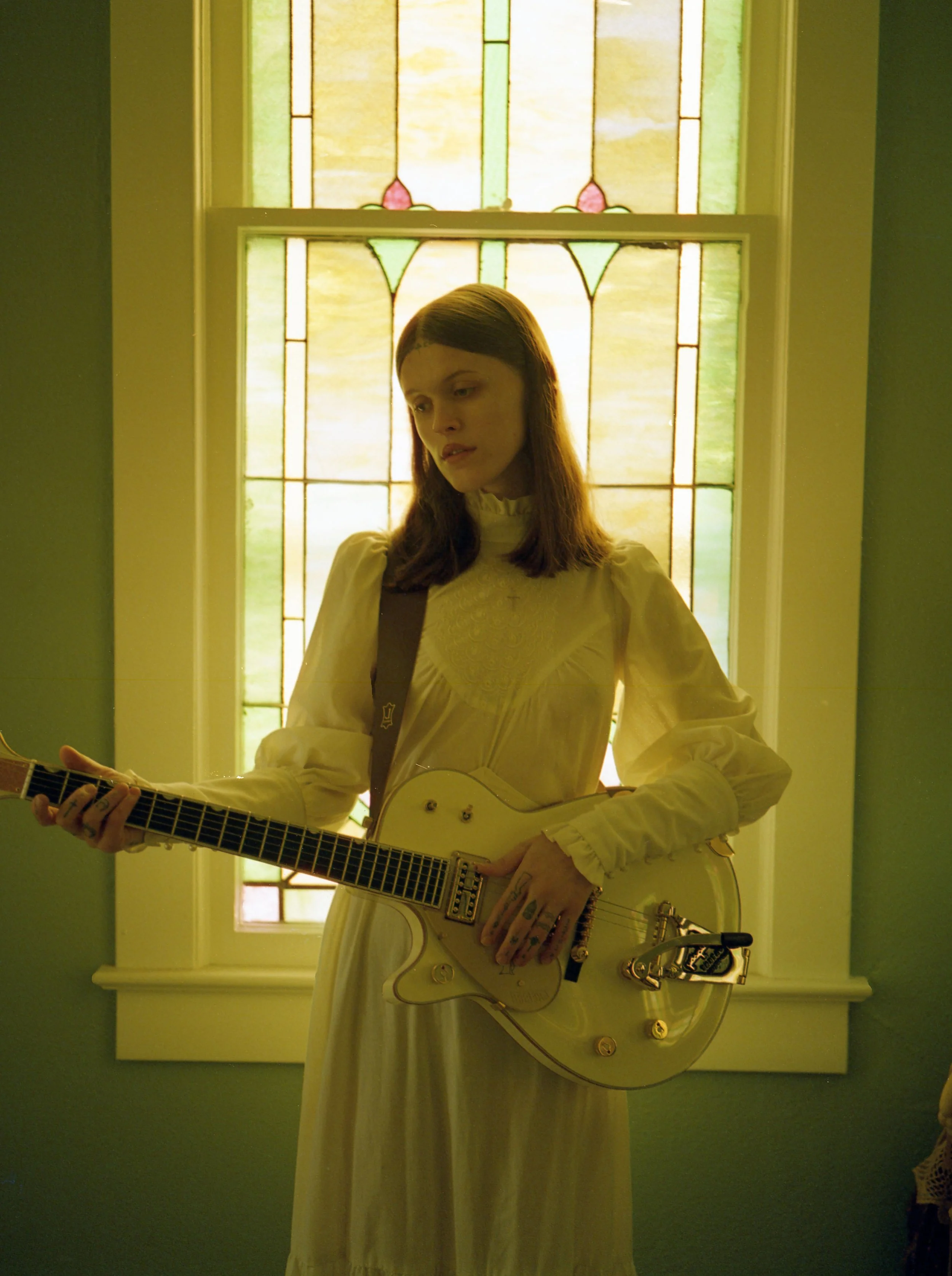 A young woman wearing a vintage white dress with long sleeves and a high collar plays an electric guitar indoors, standing in front of a stained glass window with pastel colors. Ethel Cain