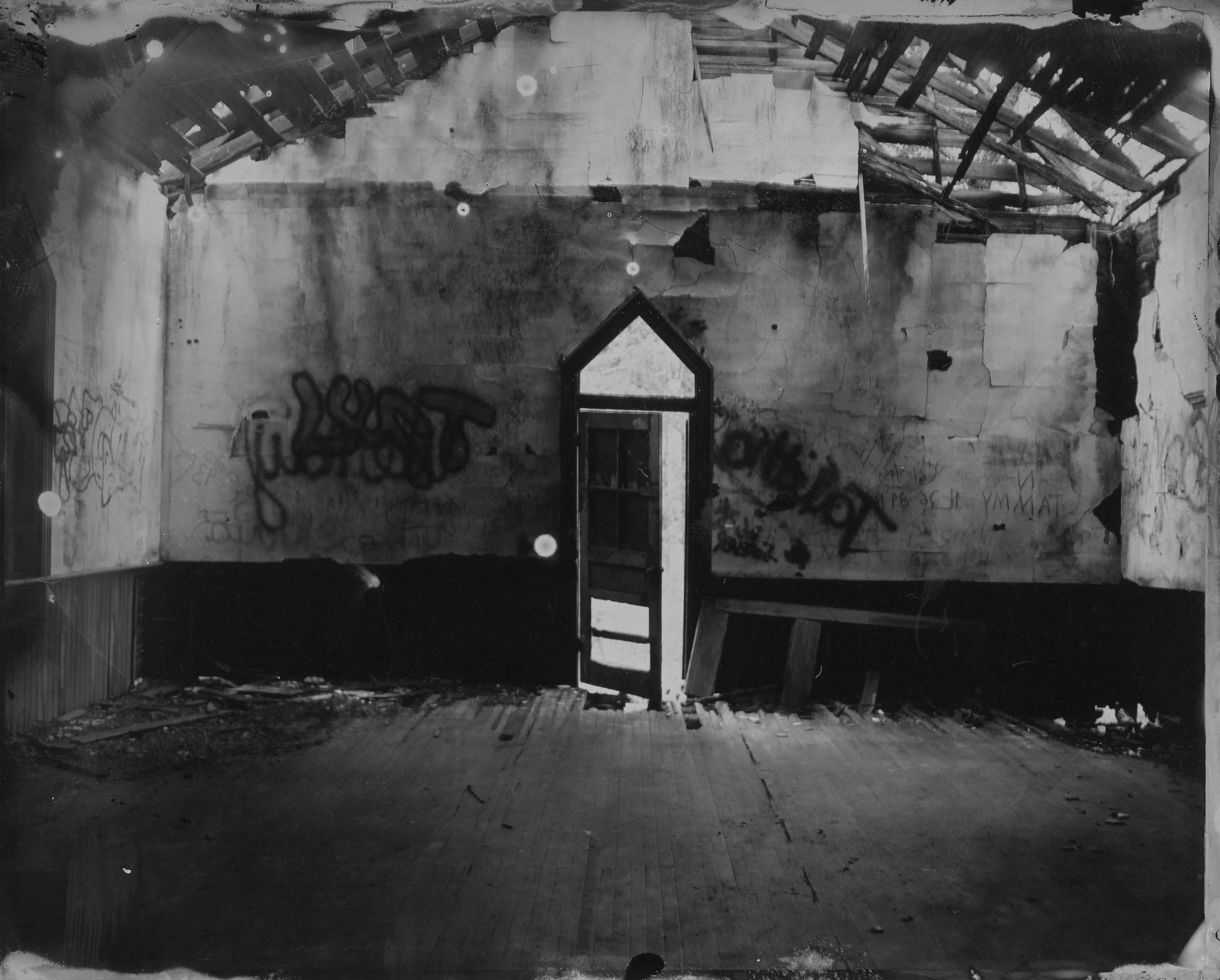 An abandoned, dilapidated room with peeling walls, graffiti, a damaged ceiling with missing shingles, and a doorway with a broken window leading outside.