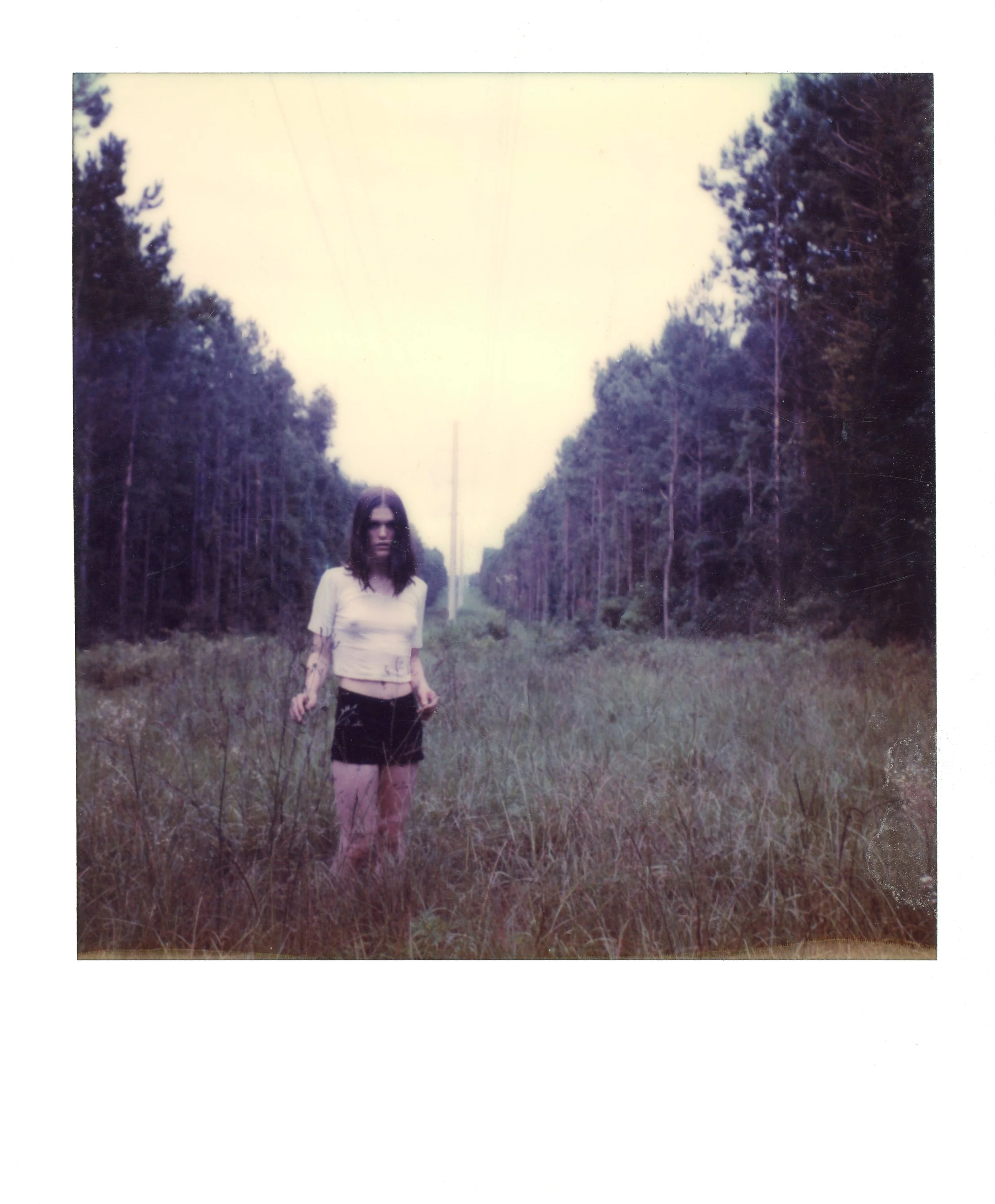 A woman with dark hair standing in a grassy field lined with tall trees, under power lines on a cloudy day.