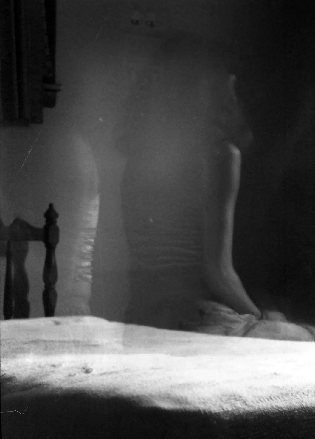A black and white photo showing a close-up of a person's bare feet standing on a bed, with a wooden headboard and dark room in the background.