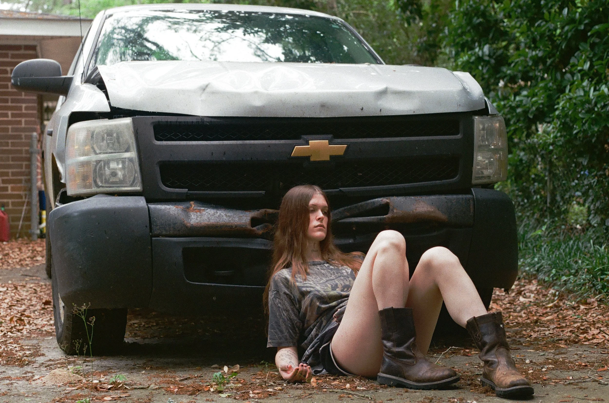 A young woman with long hair, wearing a dark t-shirt, shorts, and brown boots, sitting on the ground in front of a damaged black and white Chevrolet pickup truck with a rusty bumper Ethel Cain