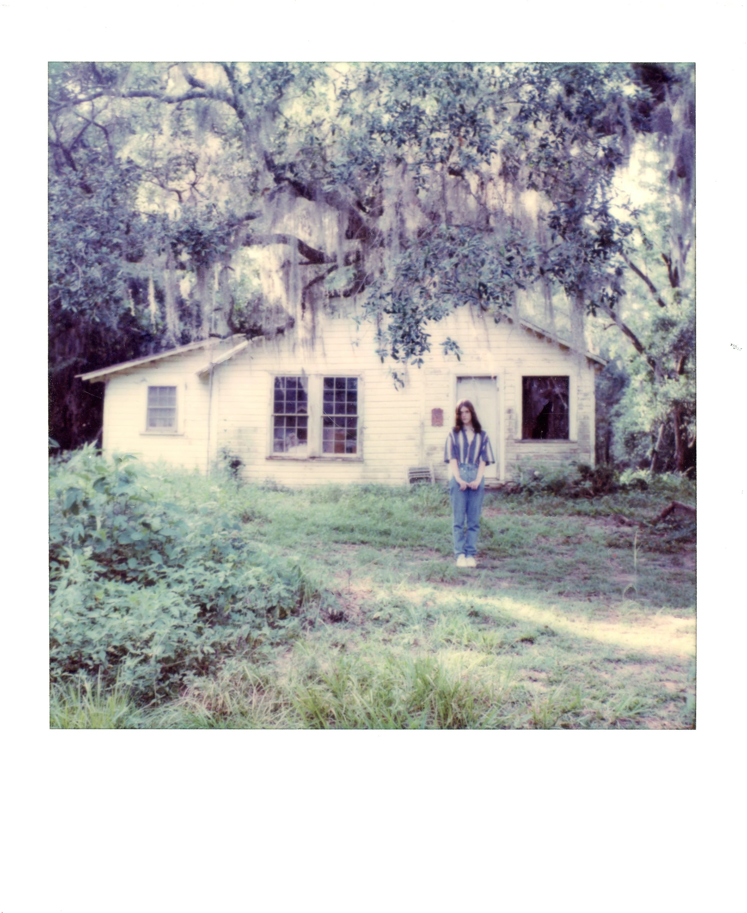 Young woman standing in front of an old house with overgrown yard and large trees overhead.