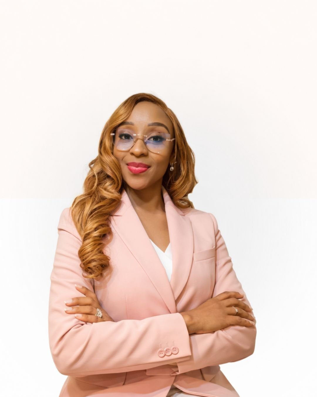 A woman with light brown, wavy hair wearing a pink blazer with crossed arms, glasses, and smiling, standing against a plain white background.