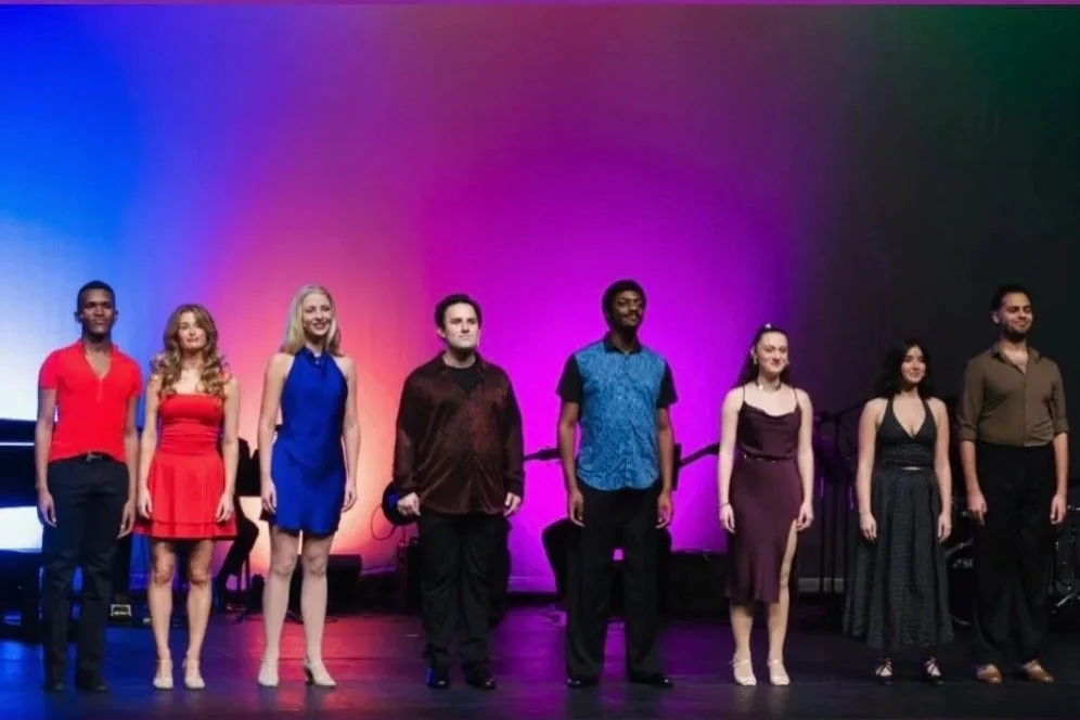 Eight performers standing on stage with colorful gradient background, some holding microphones, in formal and semi-formal attire.