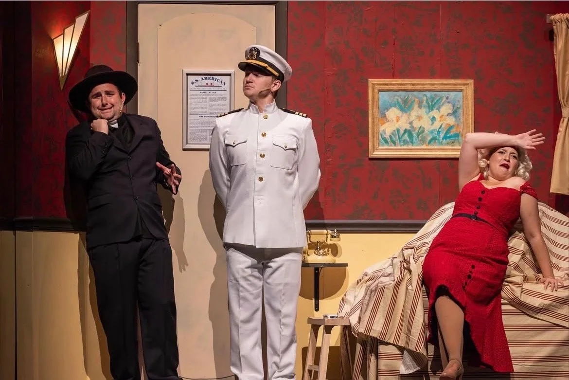 Scene from a theatrical performance with three actors on stage. One actor is dressed as a woman crying, another is dressed as a ship captain standing with hands behind his back, and the third is a woman in a red dress sitting on a bed, appearing to b