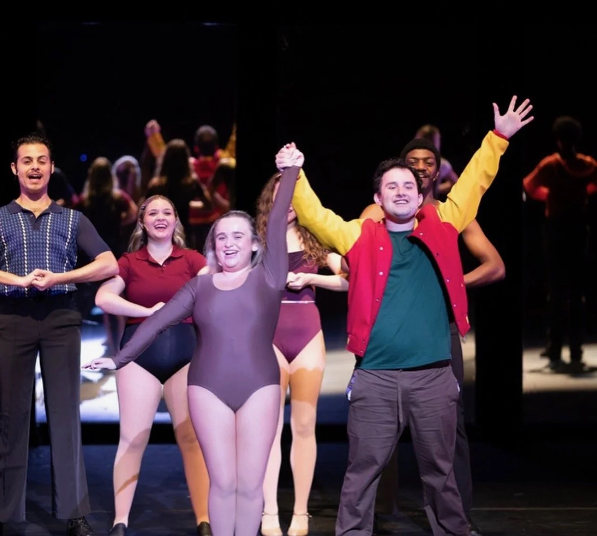 The cast of a dance or theatrical production on stage, holding hands and cheering, with some performers in colorful costumes and others in casual clothing, celebrating the show.