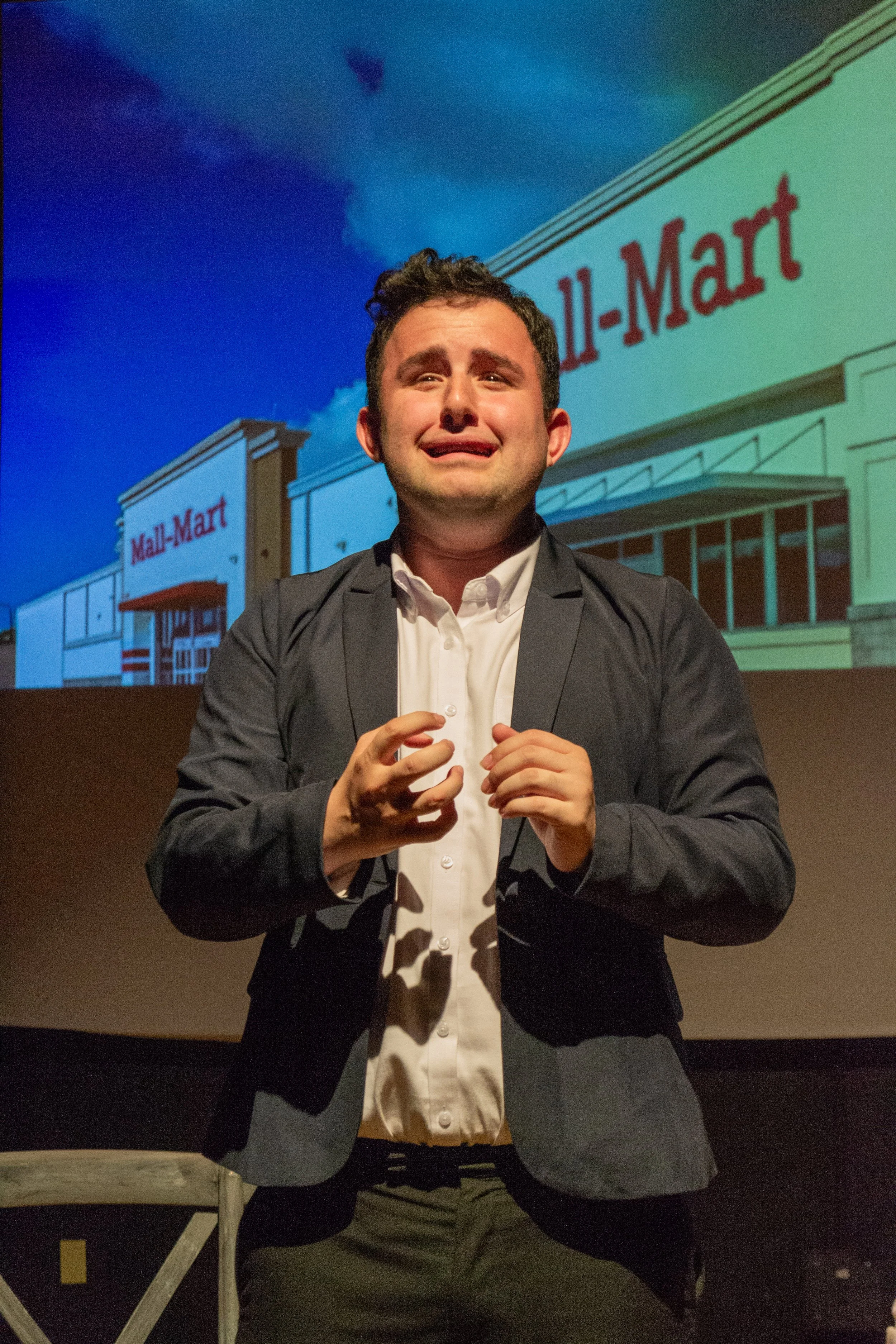 A man in a gray blazer and white shirt stands with his hands clasped in front of a Walmart building in the background. The Walmart sign is visible, and the sky behind is partly cloudy.