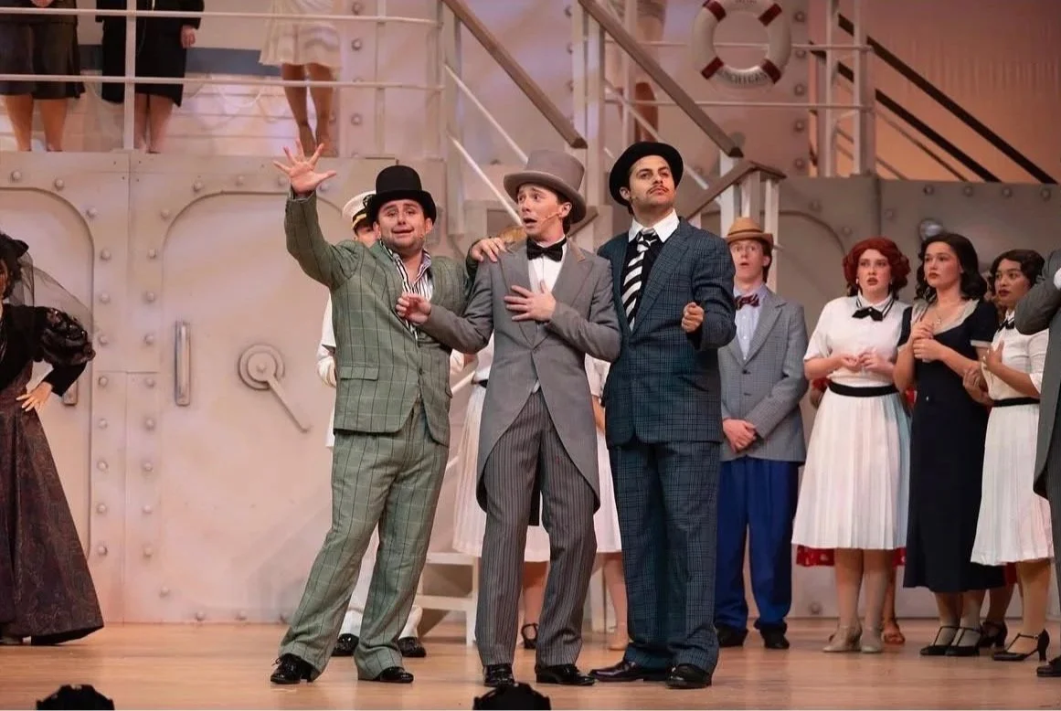 Theatrical performance on stage with actors in vintage clothing, some wearing suits and hats, portraying a scene with a mix of surprise and emotion.