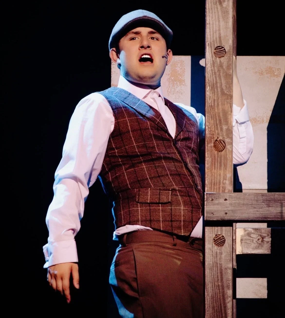 A young man is performing in a play, dressed in vintage clothing including a plaid vest, pink shirt, and a cap, standing behind a wooden structure with a black background.