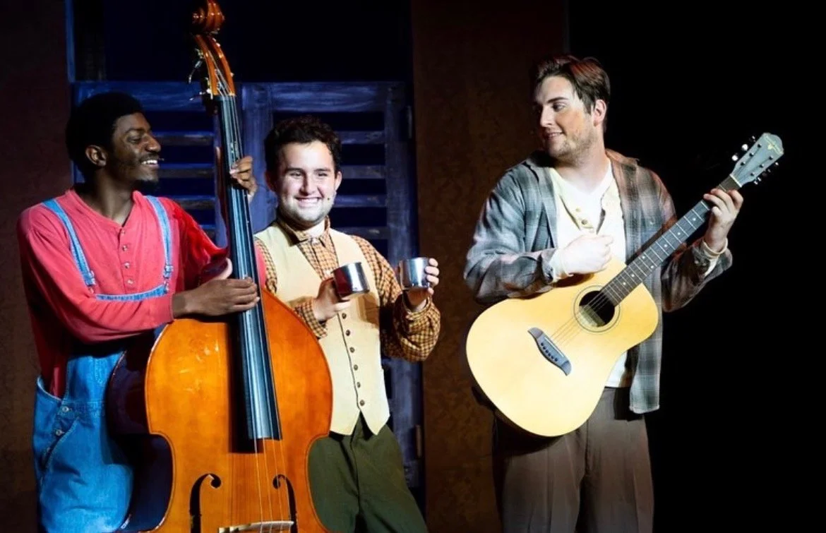 Three men playing musical instruments on stage, one with a double bass, one with a tambourine, and one with an acoustic guitar.