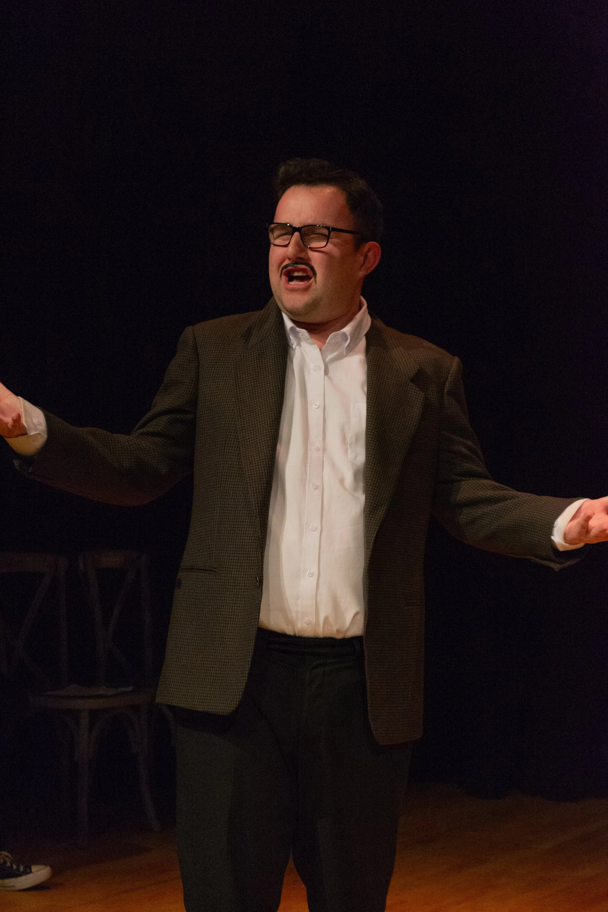 A man in glasses, a brown plaid blazer, and a white shirt appears to be singing or speaking passionately with his eyes closed, arms outstretched, against a dark background.