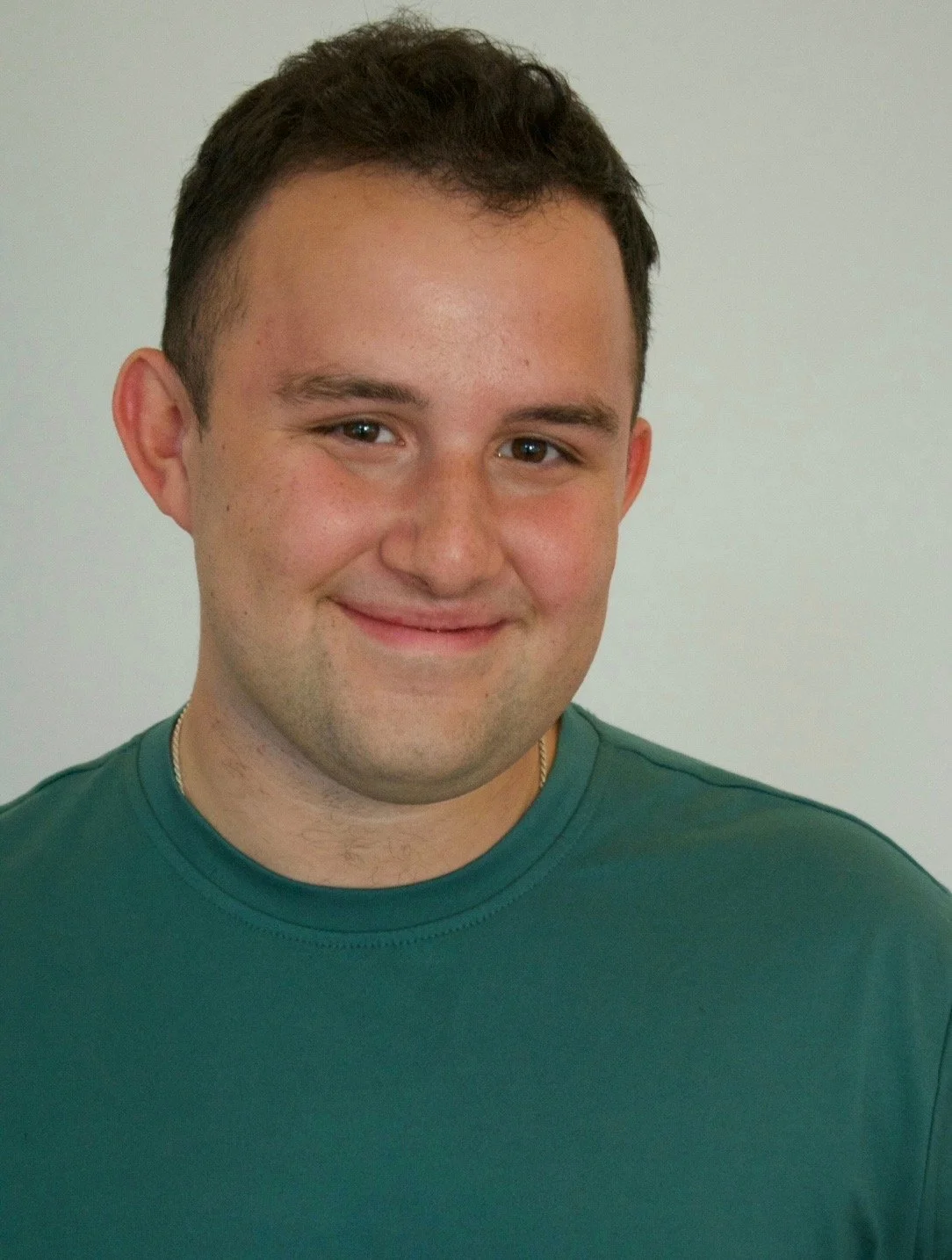 A young man with short dark hair and fair skin, smiling, wearing a green shirt, against a plain light-colored background.
