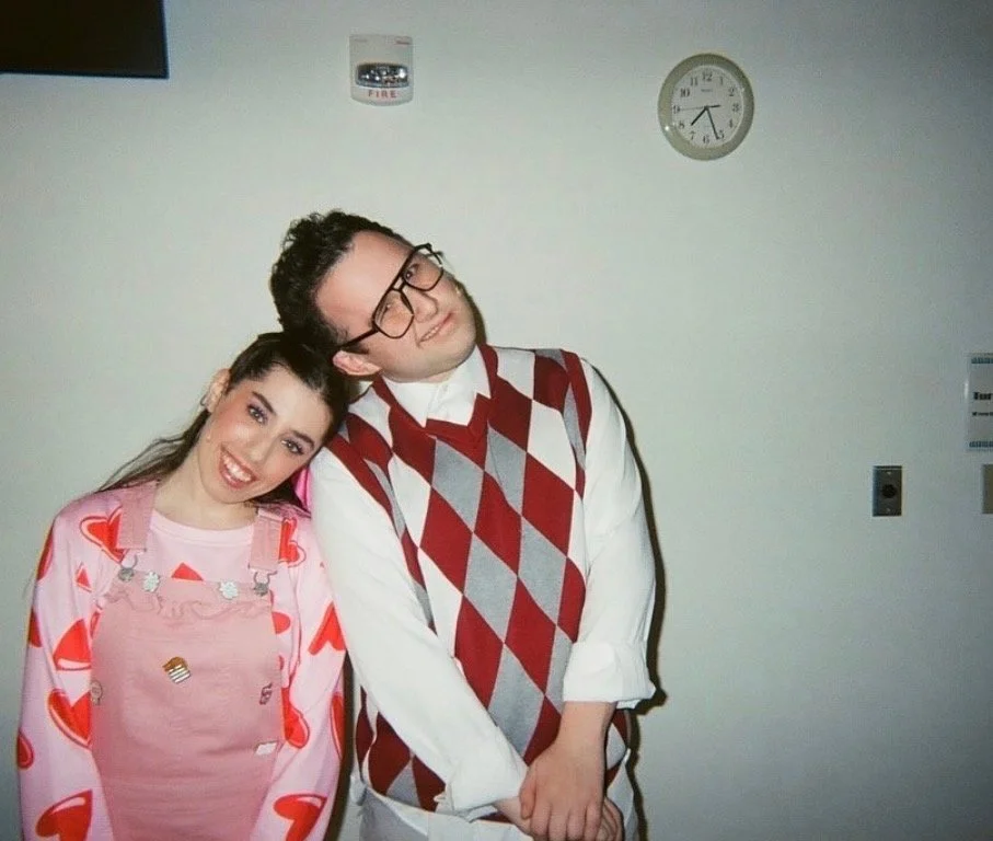 A man and woman stand close together against a plain wall. The woman is smiling and leaning her head on the man's shoulder. The woman wears a pink shirt with a heart pattern and pink overalls with various cute patches. The man has glasses, a white sh