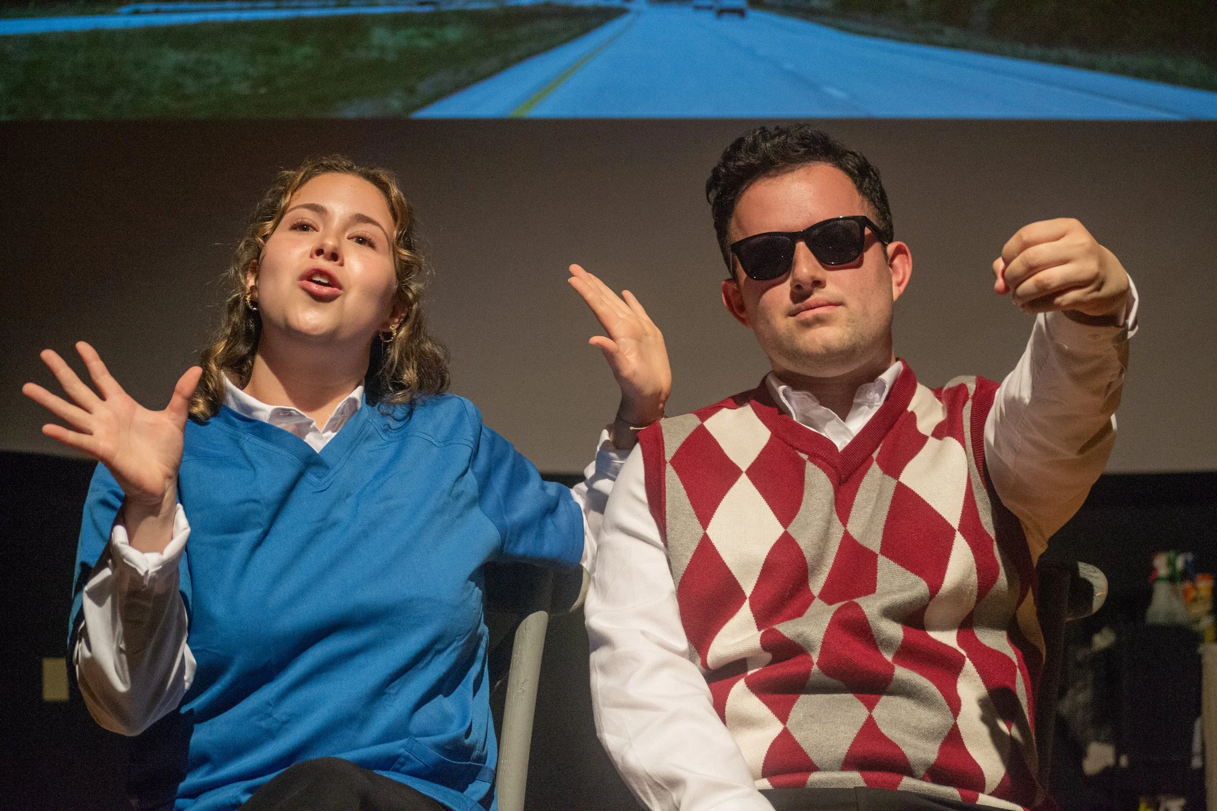 A woman wearing a blue jacket and a man with sunglasses and a red and white argyle sweater vest sitting together and gesturing outward, possibly at a presentation or event, with a dark background and a projector screen displaying a blue road scene.