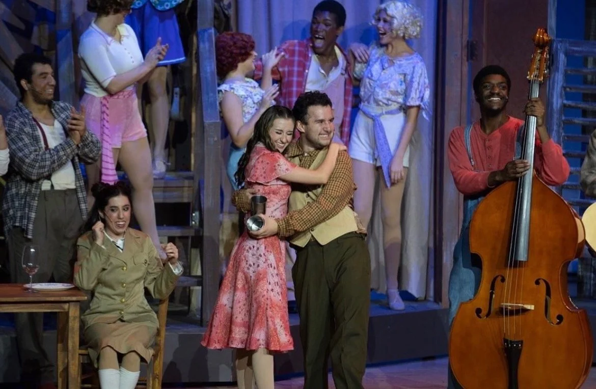 A lively group of performers on stage celebrating, with some embracing and others clapping, featuring a man playing an upright bass, and a woman sitting at a table talking on the phone.