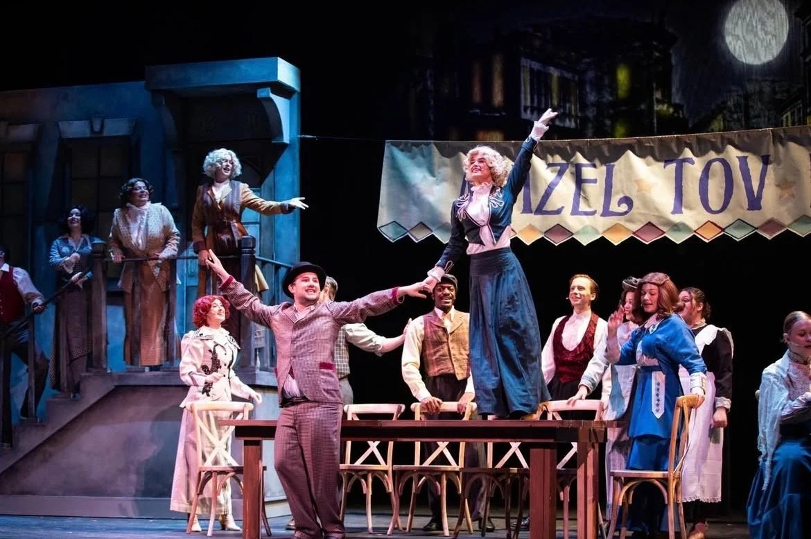 The scene appears to be from a theatrical performance, with actors on stage dressed in period costumes. Two performers are in the center, with one standing on a table and the other holding her hand, smiling and engaging with each other. Additional ca